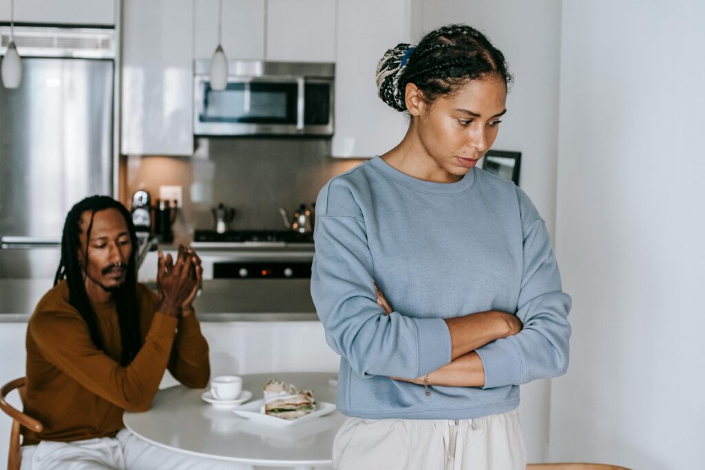 Young discontented African American female with folded arms against male partner at kitchen table during quarrel